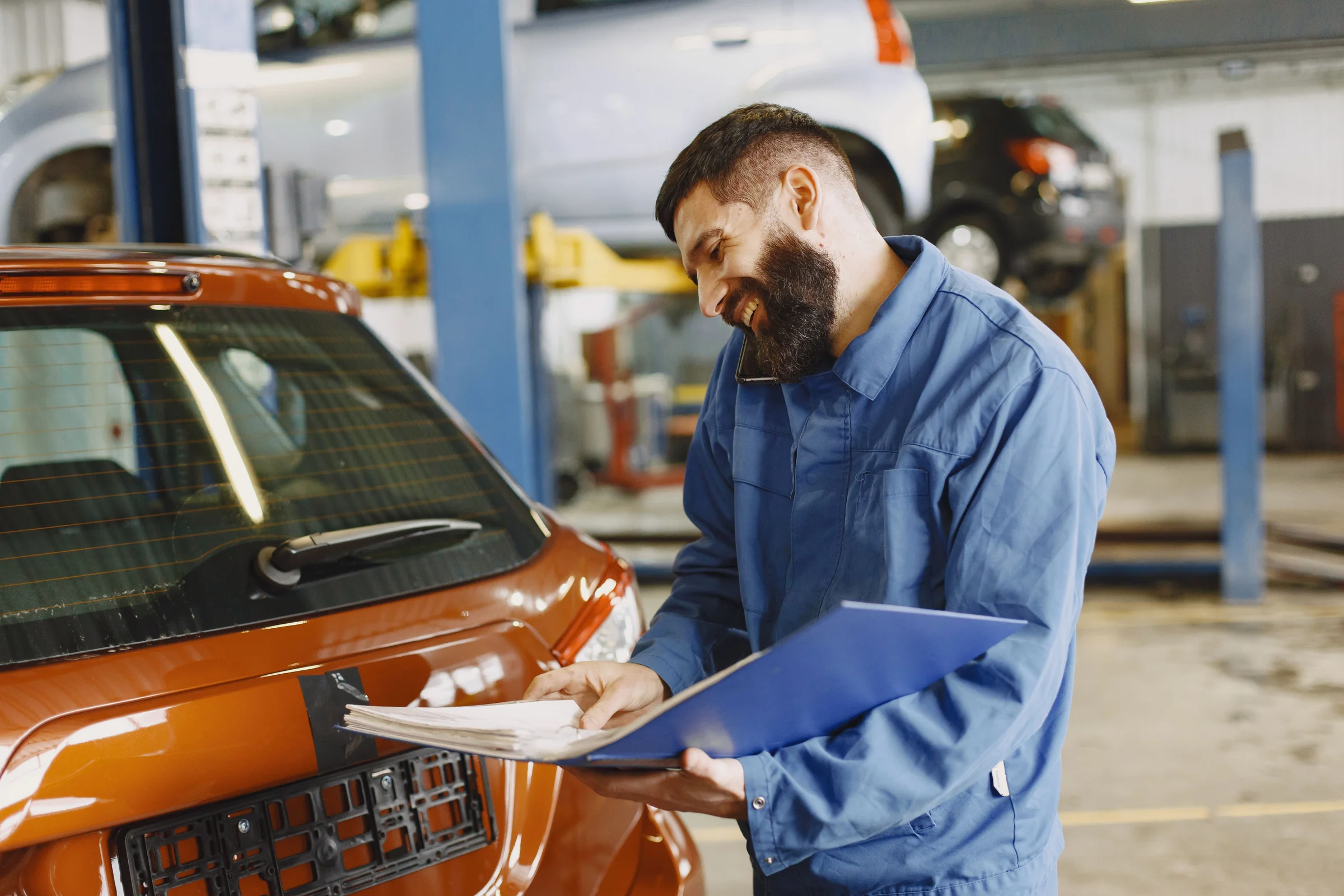 A mechanic in a blue work uniform noting details on a clipboard while inspecting an orange car in a service garage